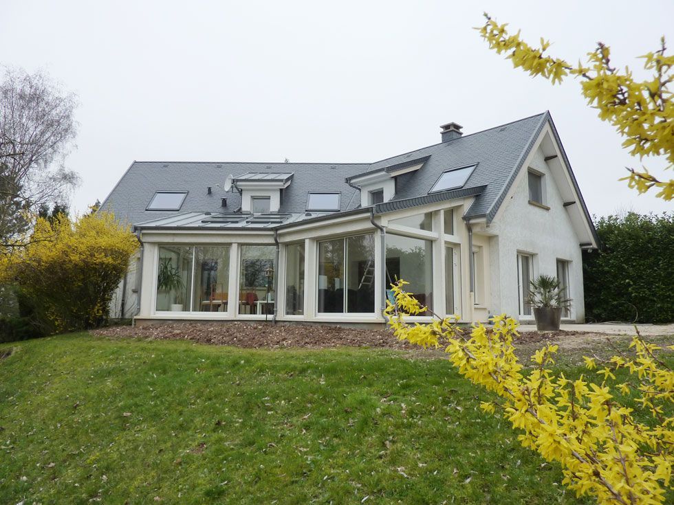 Maison blanche avec véranda vitrée, toit gris, fleurs jaunes et pelouse verte.