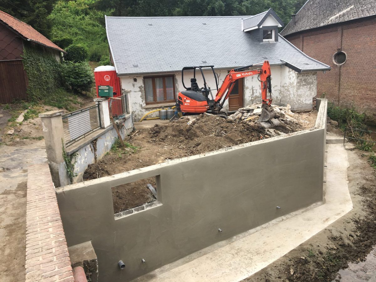 Chantier de construction avec une petite excavatrice, un bâtiment et un mur fraîchement enduit.