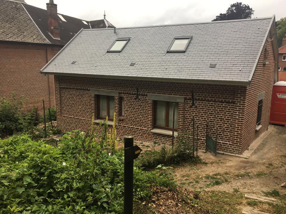 Bâtiment en briques avec deux puits de lumière, deux fenêtres et un toit gris, situé dans une cour verdoyante.