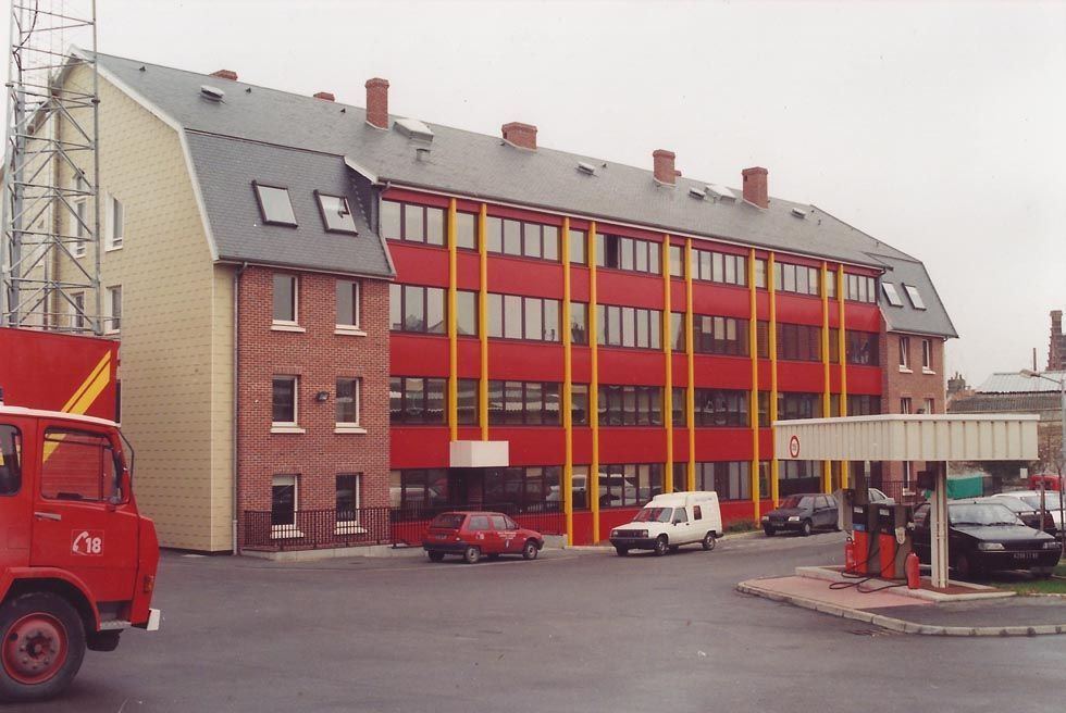 Caserne de pompiers à façade rouge et supports jaunes ; camion de pompiers au premier plan.
