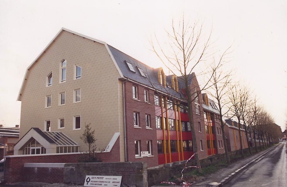 Immeuble en briques à plusieurs étages avec des touches de beige et de rouge, près d'une route bordée d'arbres dénudés.