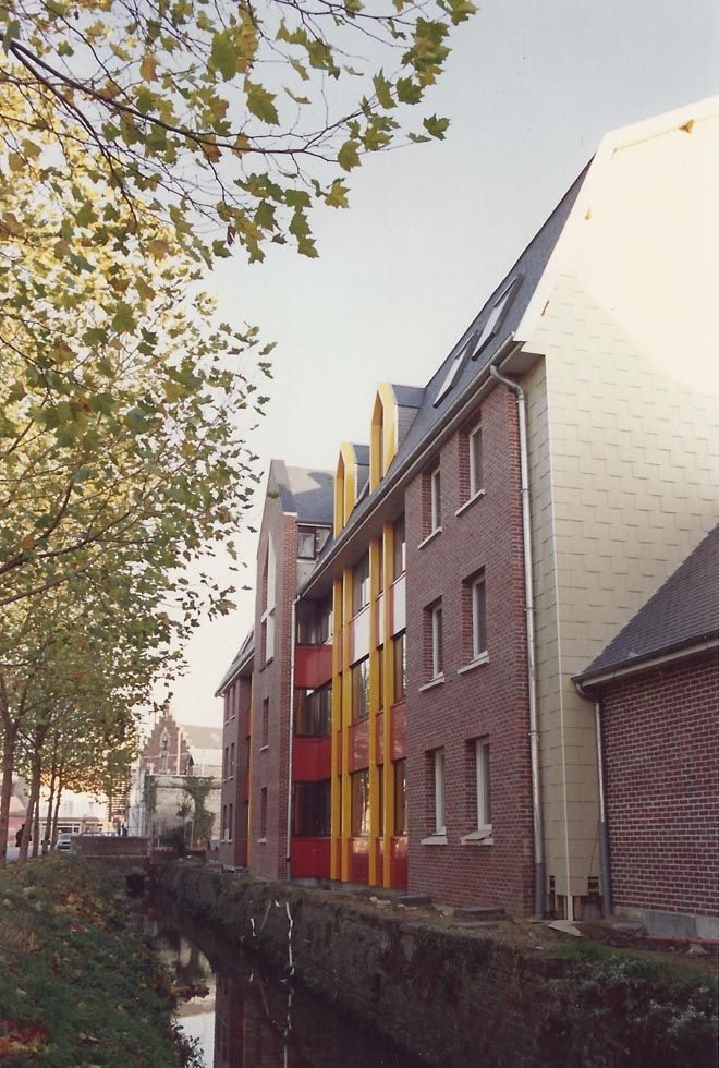 Bâtiments en briques bordant un canal, avec des touches de jaune et de rouge. Arbres au premier plan.