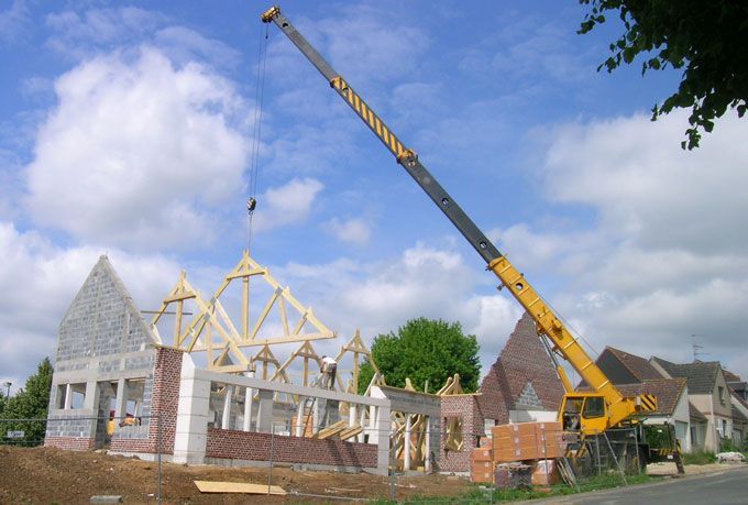 Chantier de construction : une grue soulève des poutres en bois pour une maison aux murs de briques et de pierres, sous un ciel bleu nuageux.