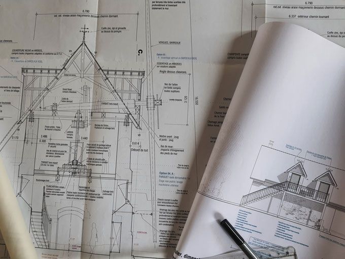 Plans architecturaux d'un bâtiment à toit pointu, croix et schémas. Un rouleau de papier et un stylo se trouvent à proximité.