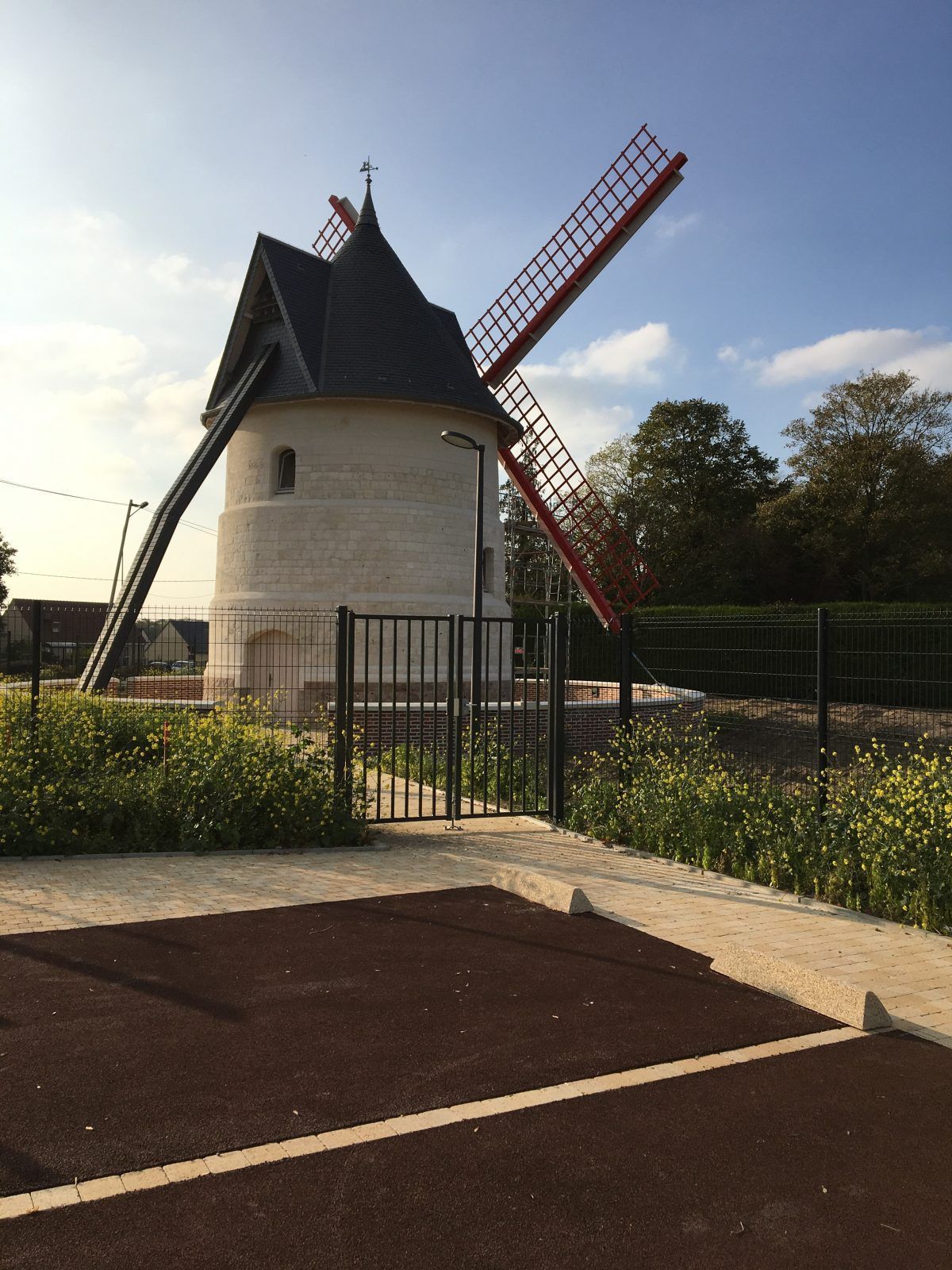 Moulin à vent blanc aux ailes rouges sous un ciel bleu, entouré d'une clôture et de verdure.