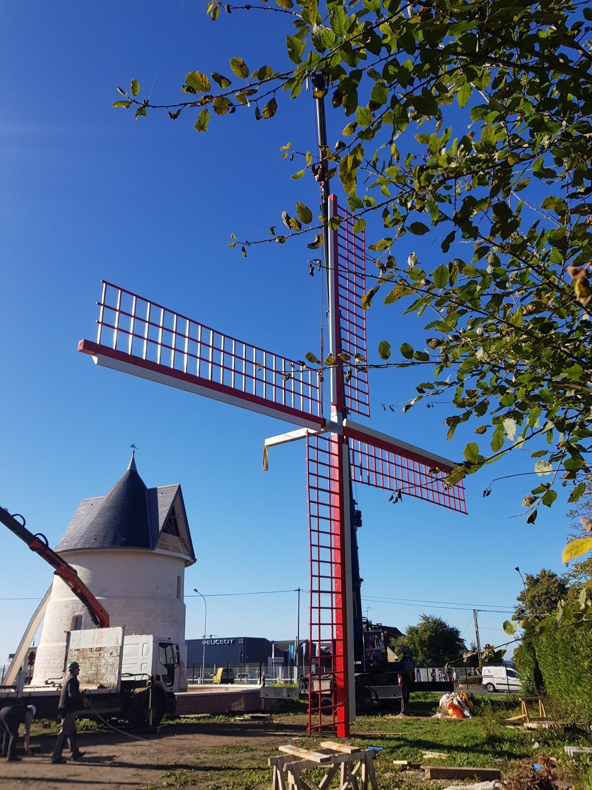 Par une journée ensoleillée, on construit un moulin à vent aux pales rouges et blanches. Un bâtiment au toit conique se dresse à l'arrière-plan.
