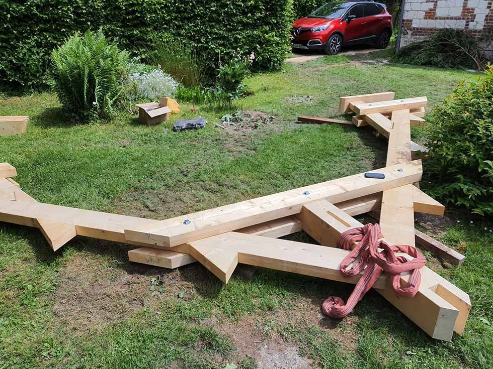 Poutres et supports en bois sur l'herbe d'une cour, près d'une voiture rouge et de buissons.