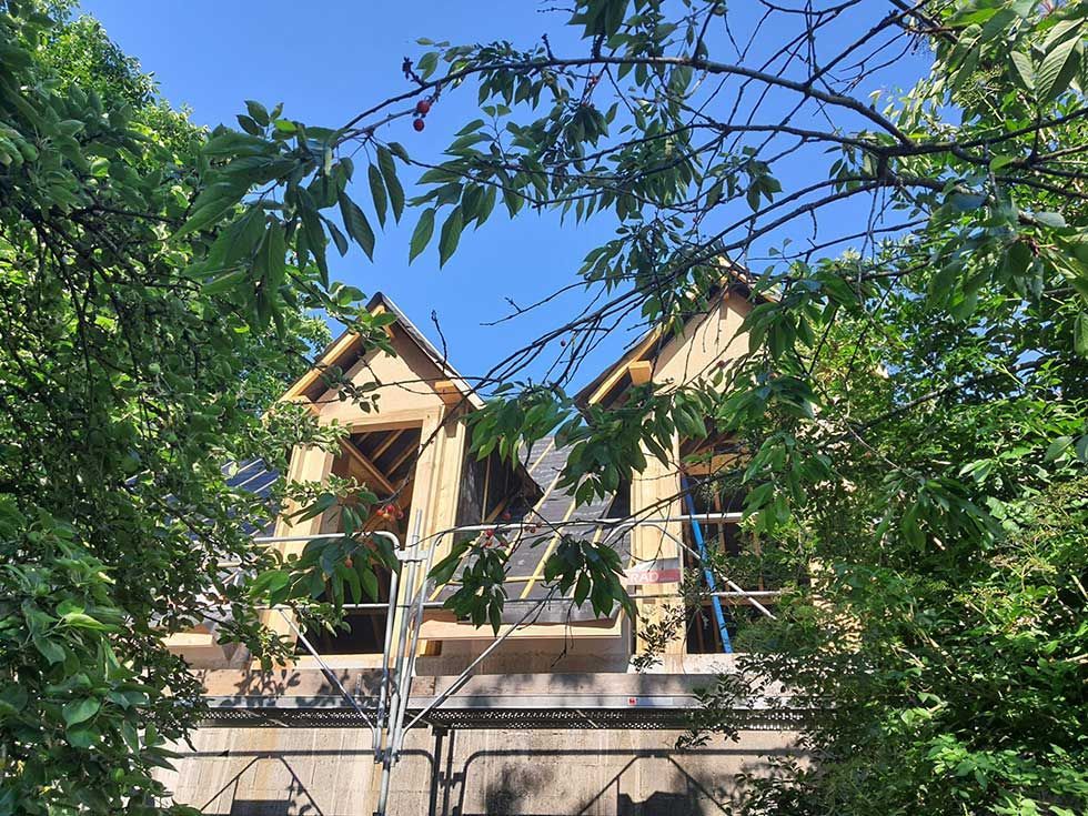 Deux lucarnes en construction, aux charpentes apparentes, que l'on aperçoit à travers les branches d'un arbre sur fond de ciel bleu.