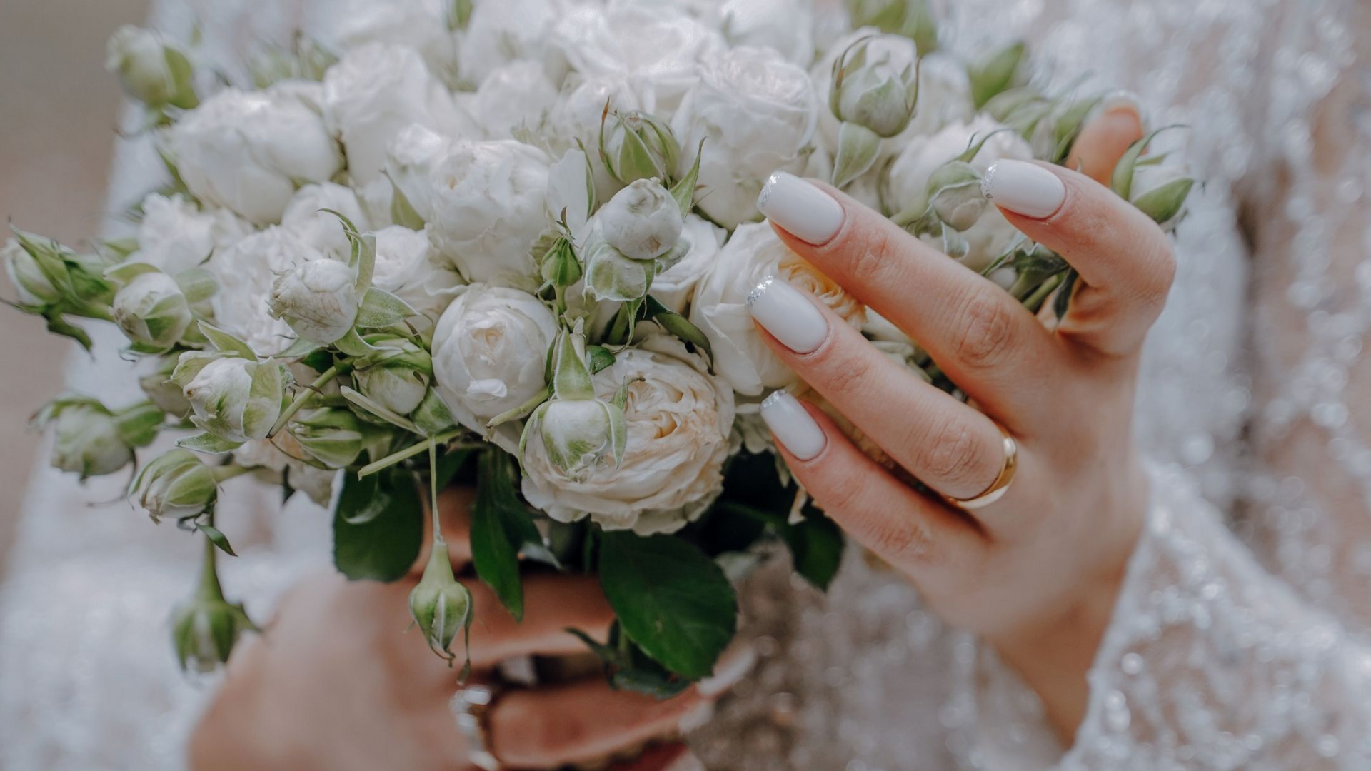 Bouquet de mariée blanc composé de roses et de verdure, tenu par des mains aux ongles blancs manucurés et à la manche en dentelle