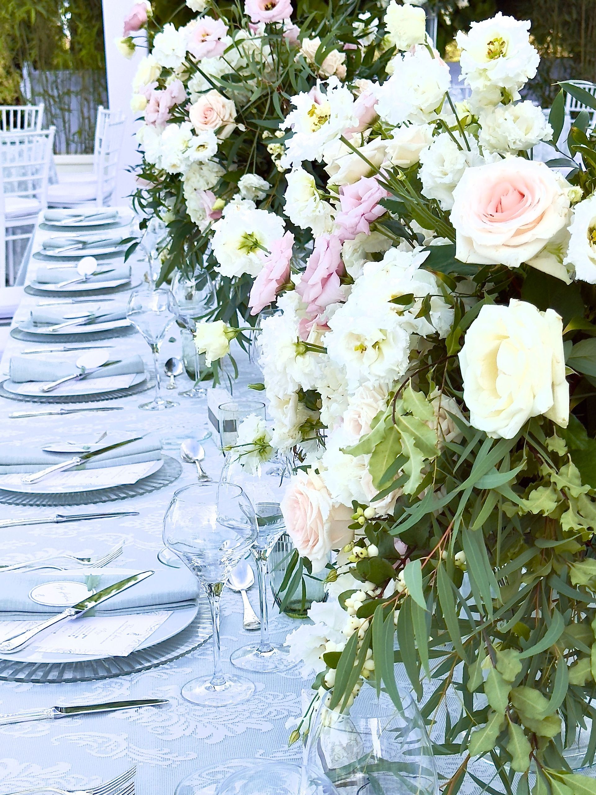 Élégant chemin de table fleuri blanc et rose poudré à côté d'une table dressée avec de la verrerie et des assiettes.