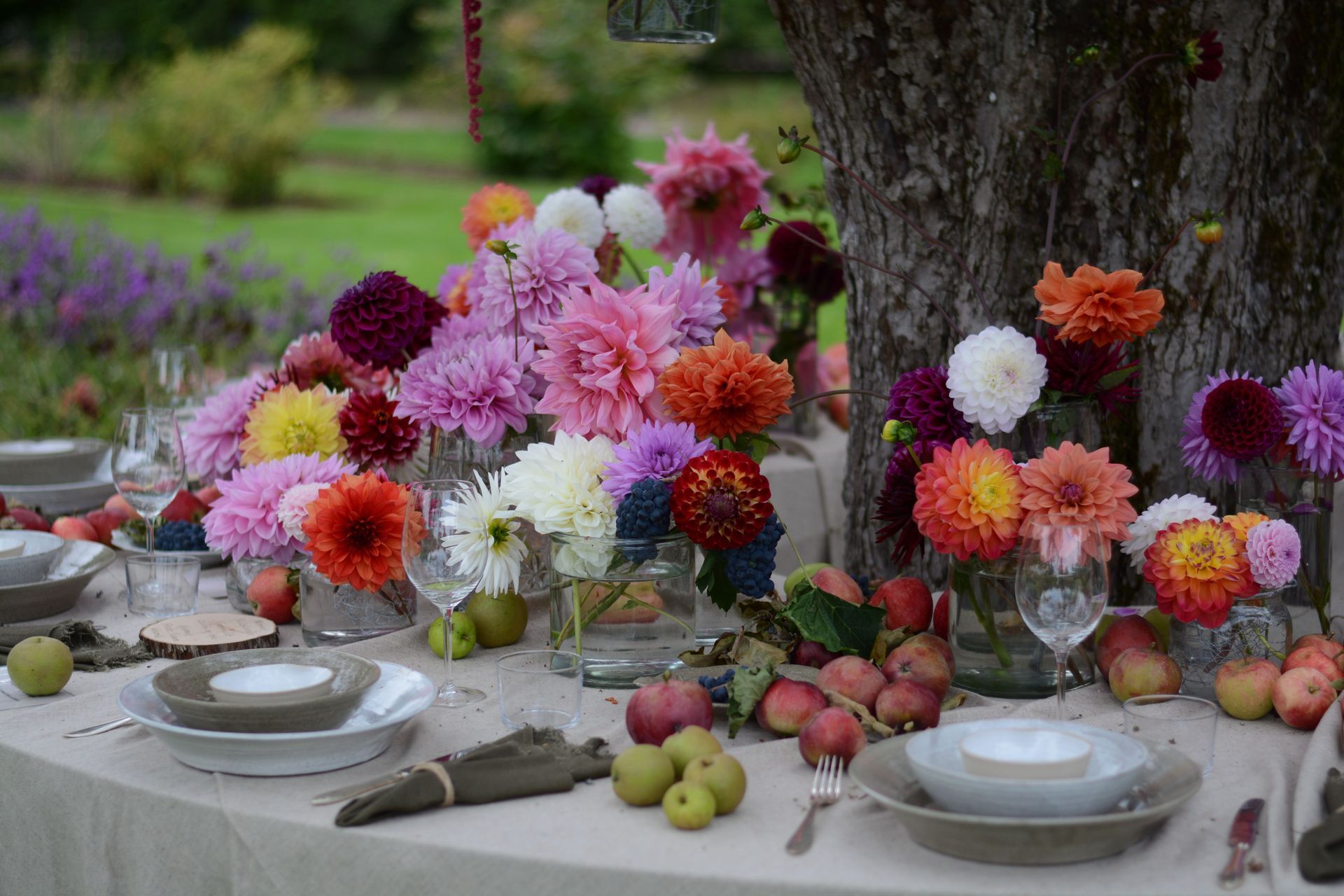 Table d'extérieur dressée avec des compositions florales colorées, de la vaisselle blanche et des fruits sous un arbre