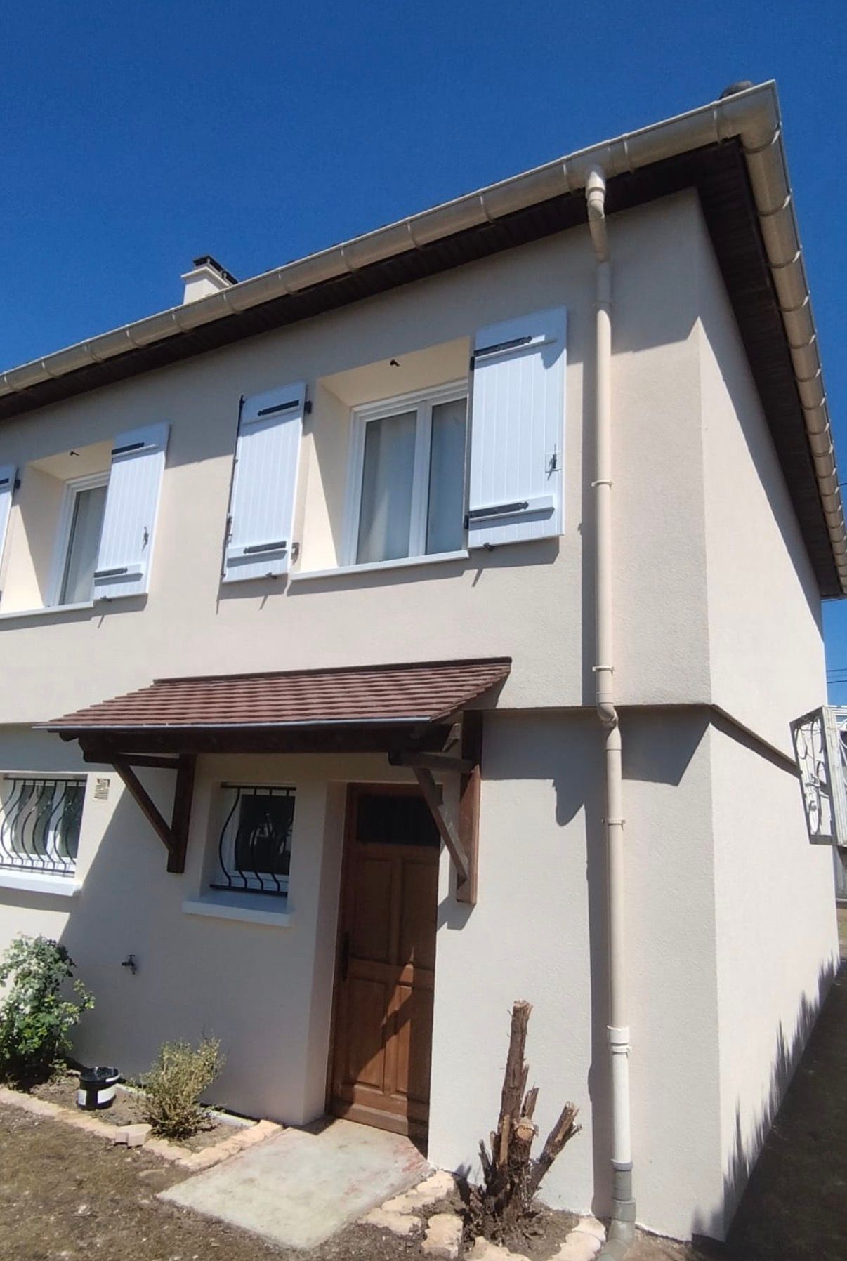 Maison beige à deux étages avec des volets blancs, une porte marron et un petit toit en porche, sous un ciel bleu.