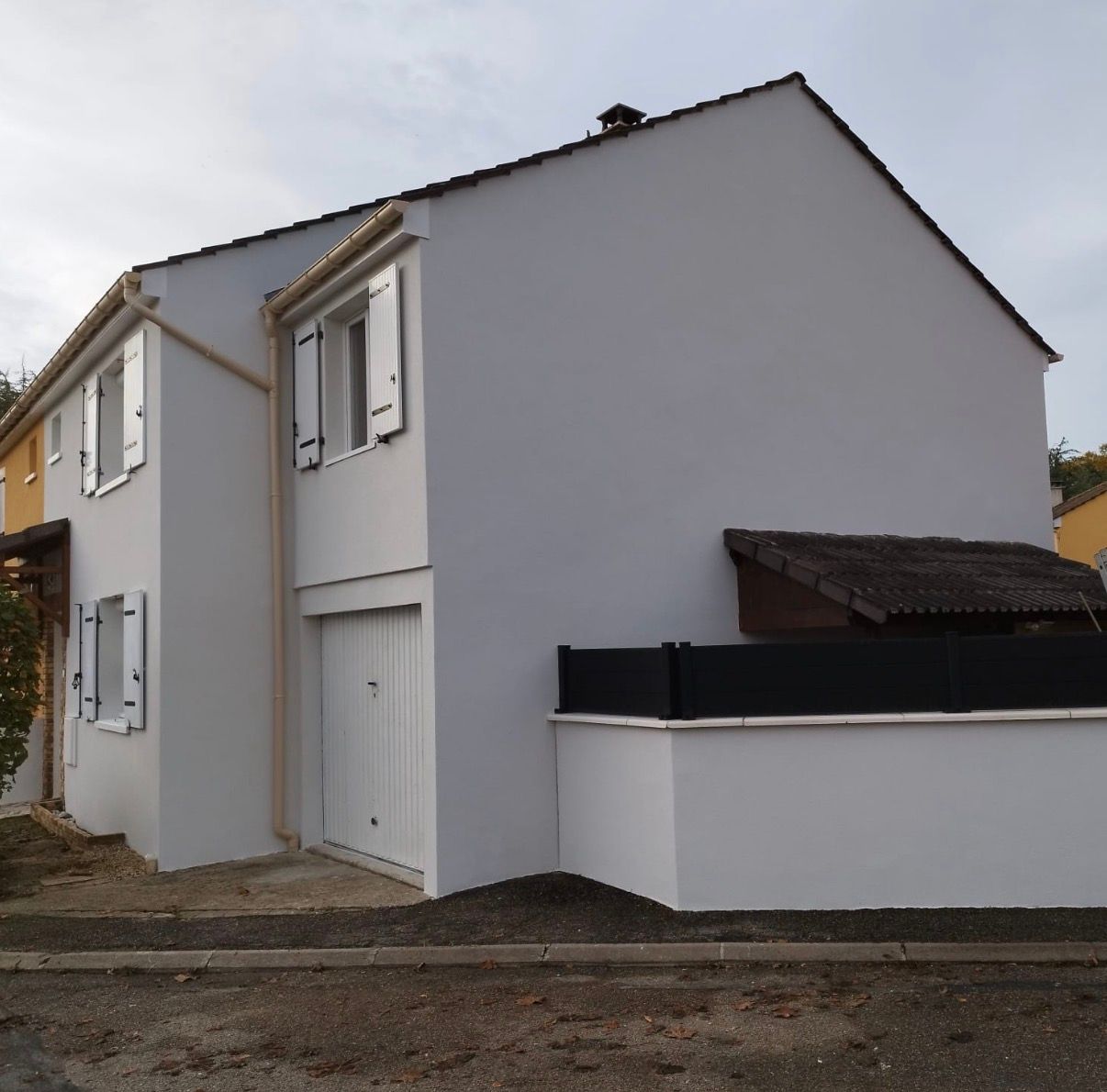 Maison à deux étages avec murs gris clair, volets et porte de garage blancs. Clôture et toit noirs visibles.