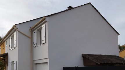 Maison à deux étages avec murs gris clair, toit foncé, volets blancs et gouttières beiges.