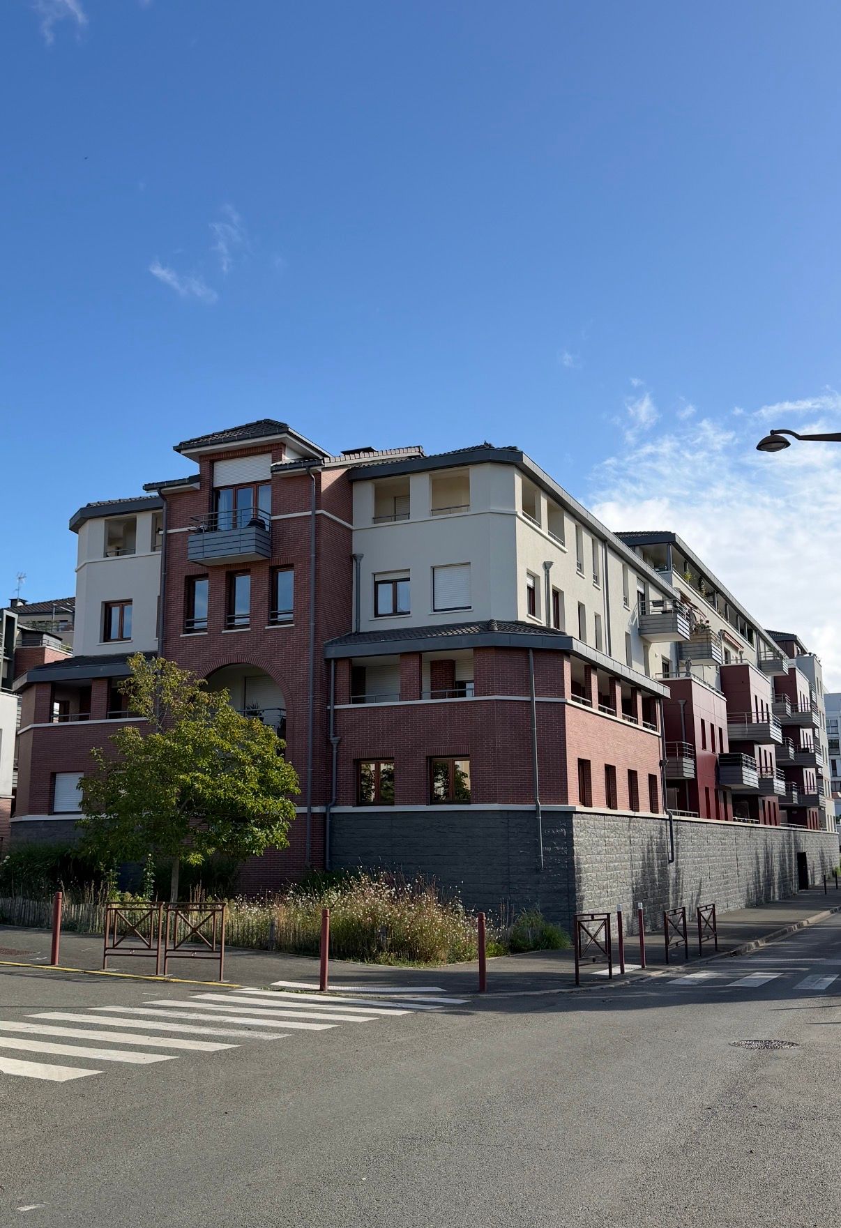 Immeuble d'habitation en briques rouges et murs beiges, sur fond de ciel bleu. Un petit arbre et un passage piéton se trouvent devant.
