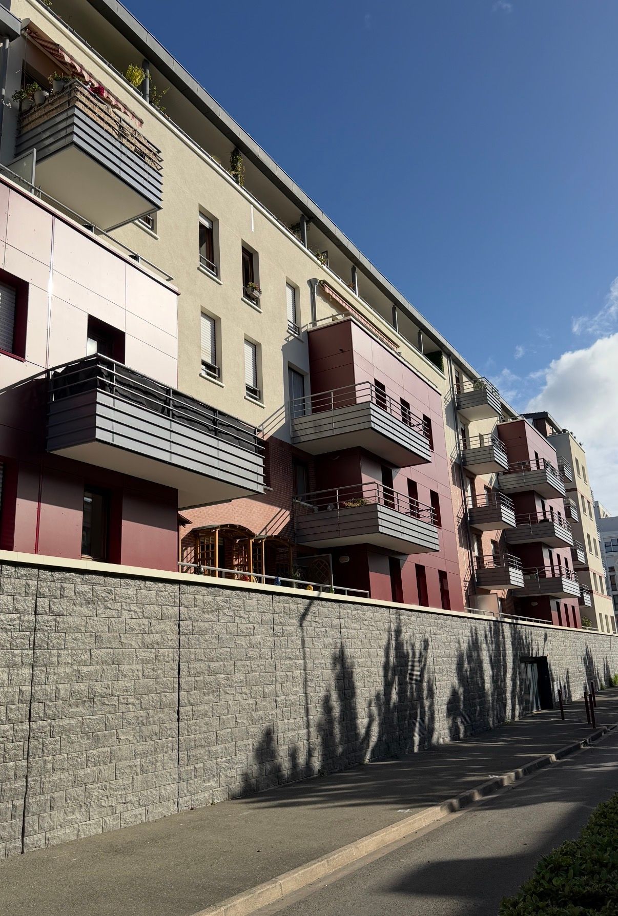Immeuble à plusieurs étages avec balcons, mur en pierre grise, ciel bleu.