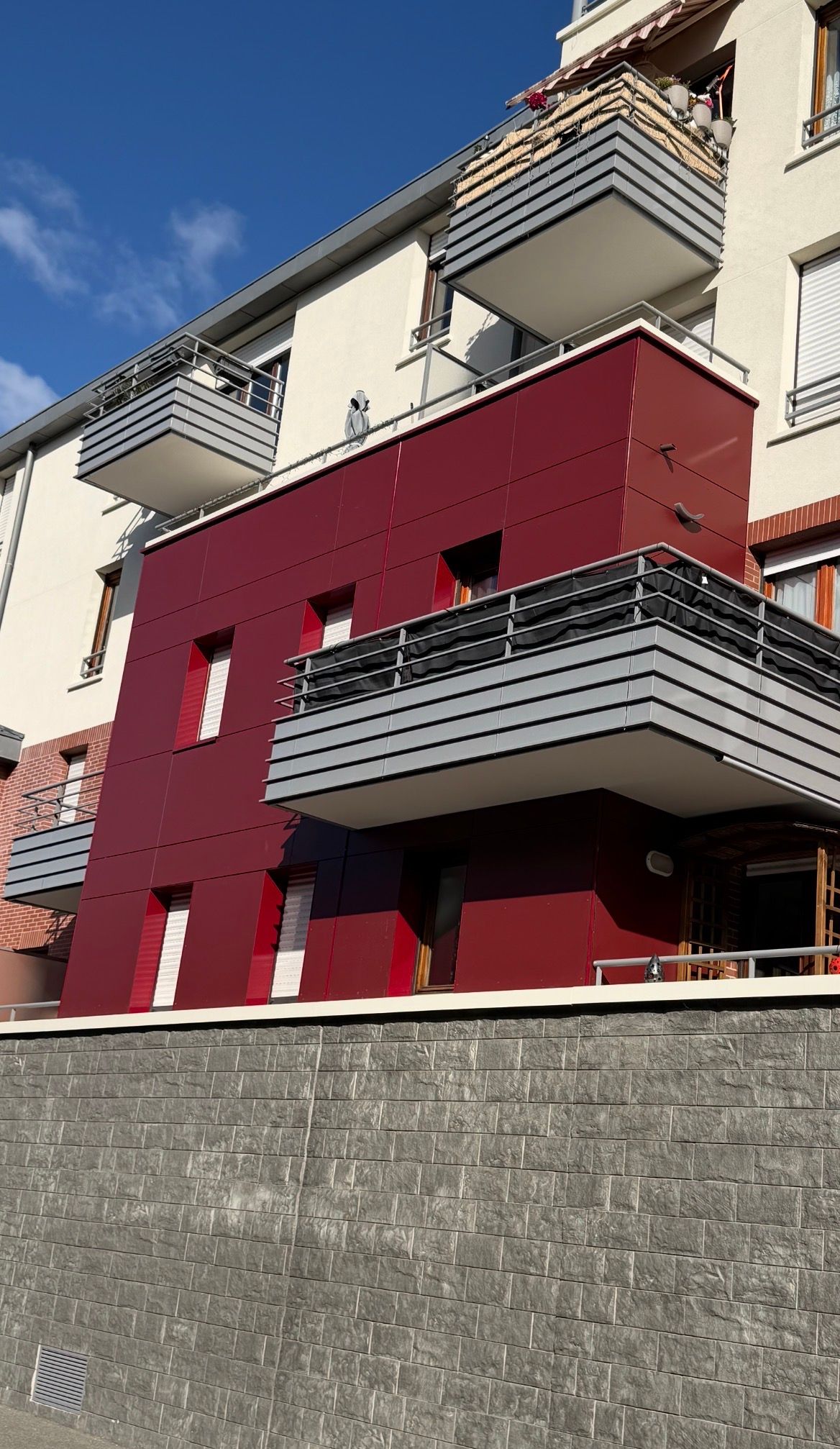 Bâtiment aux murs rouges avec balcons attachés à un immeuble à plusieurs étages contre un ciel bleu.