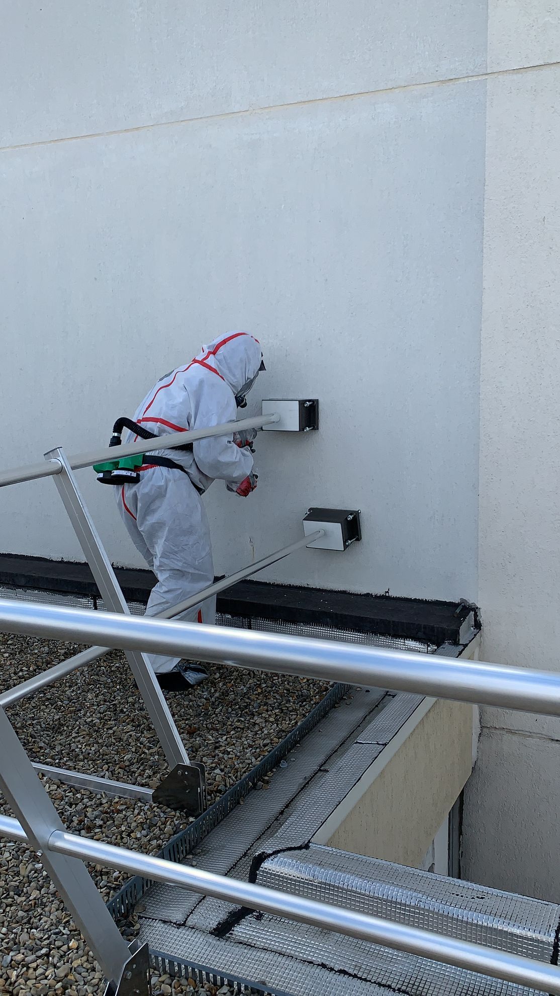 Personne en combinaison de protection travaillant sur un mur, à l'aide d'outils, à côté d'une balustrade métallique.
