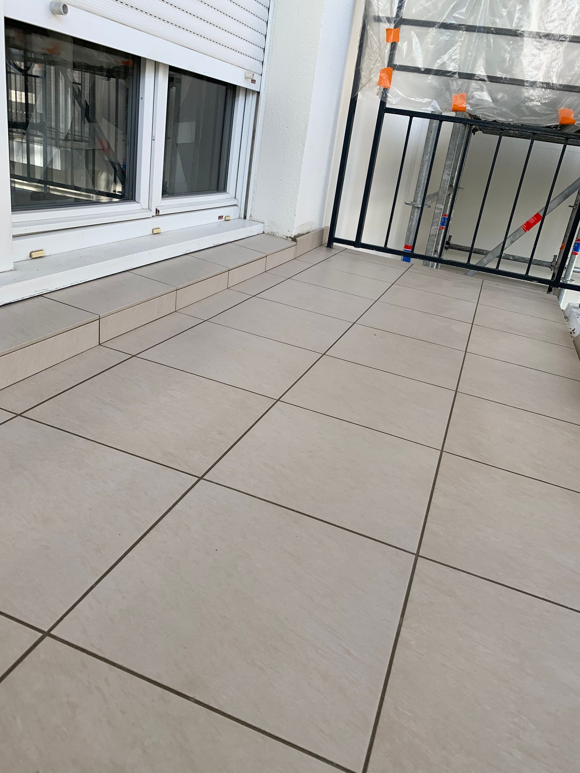 Balcon avec sol carrelé de couleur claire, garde-corps en métal noir et fenêtre à cadre blanc avec volets.