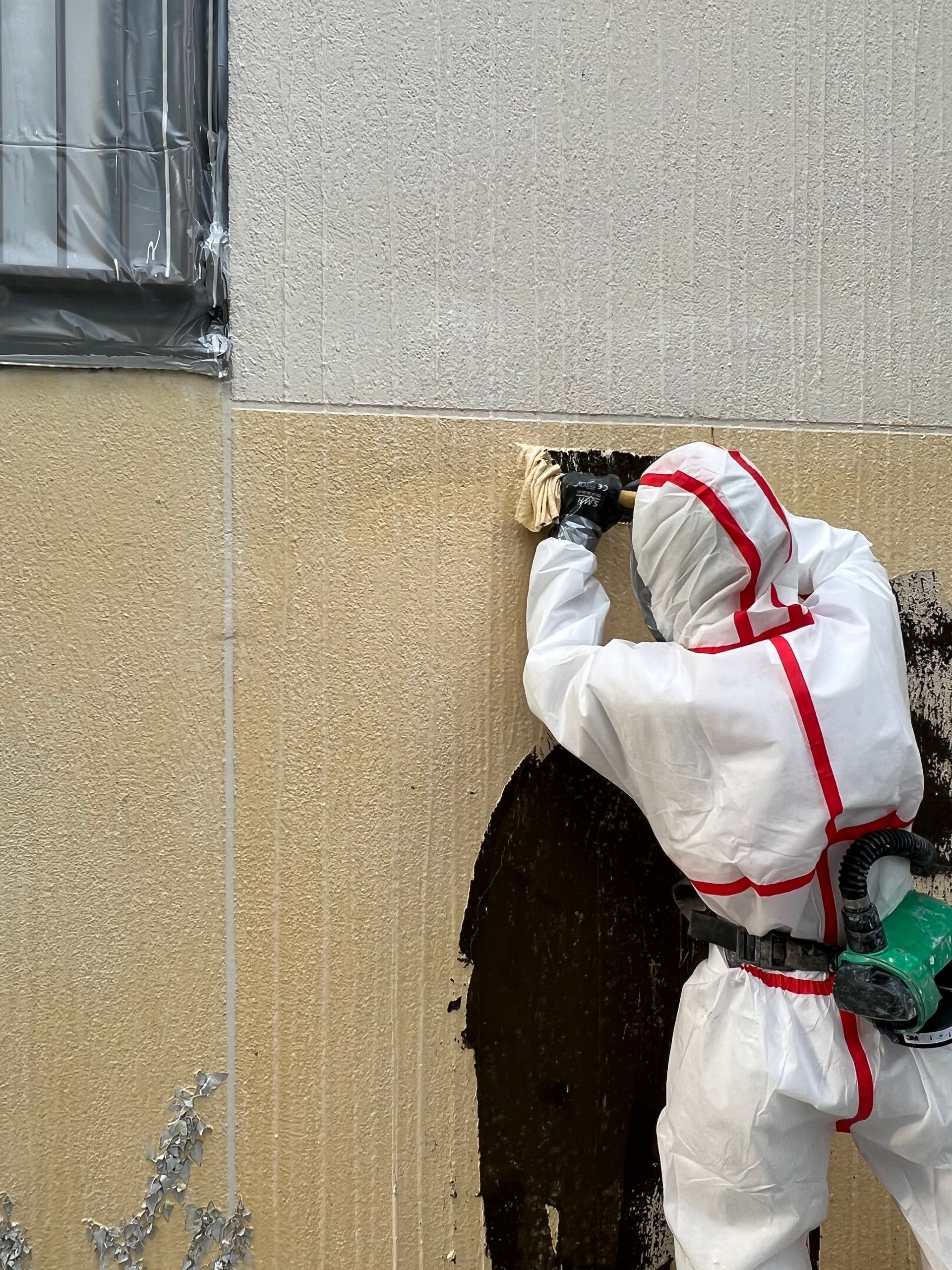 Une personne portant une combinaison de protection blanche retire des matériaux de l'extérieur du bâtiment à l'aide d'un outil électrique.