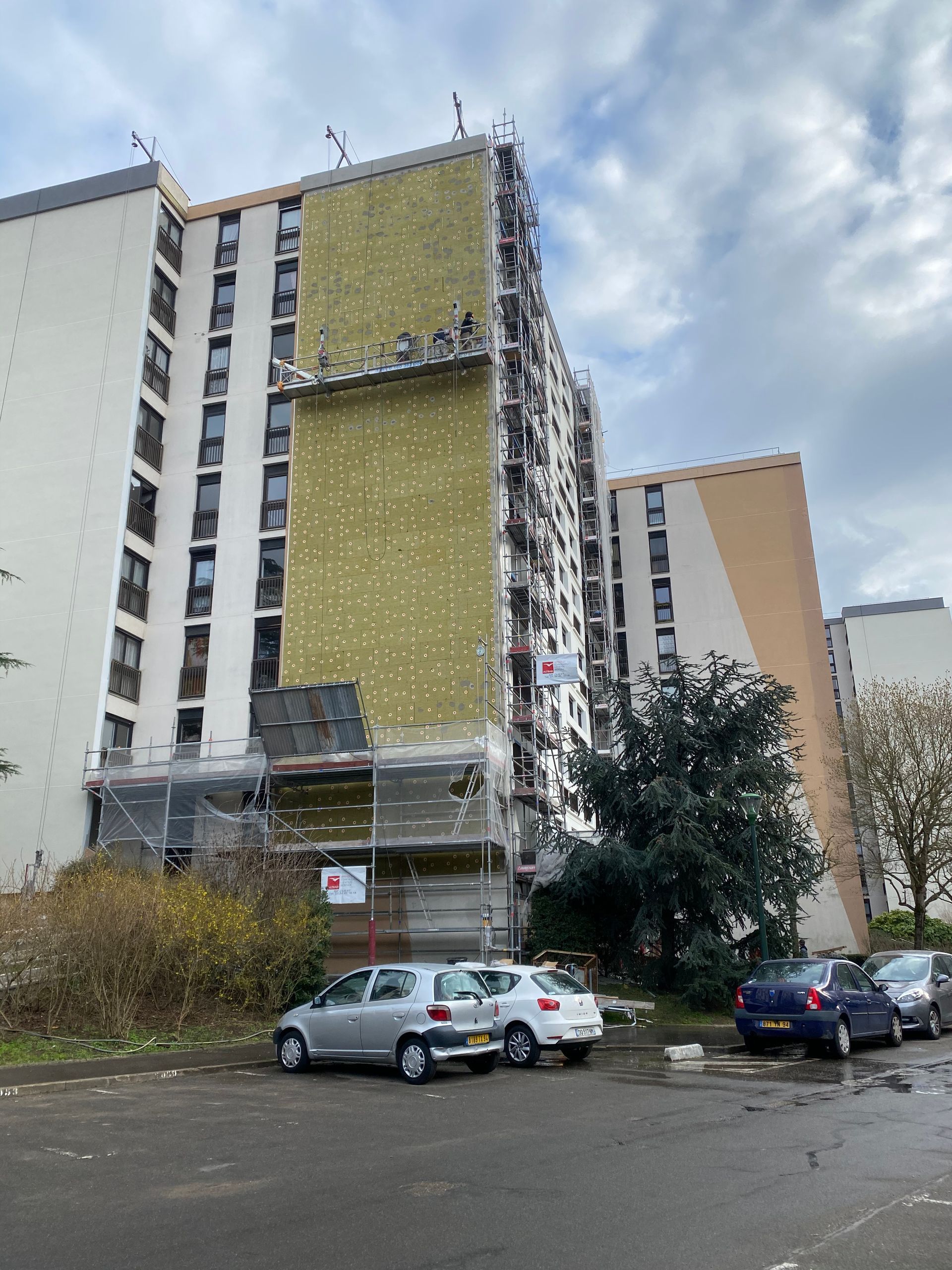 Immeuble de plusieurs étages en cours de rénovation ; échafaudages et isolation visibles. Voitures garées au premier plan.