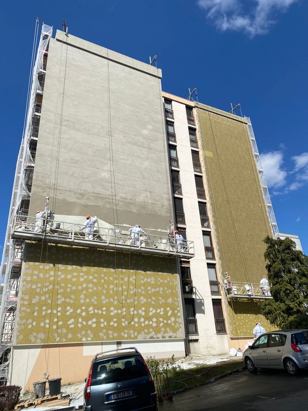 Immeuble à plusieurs étages avec échafaudages. Mur extérieur en cours de rénovation et d'isolation ; voitures garées en contrebas.