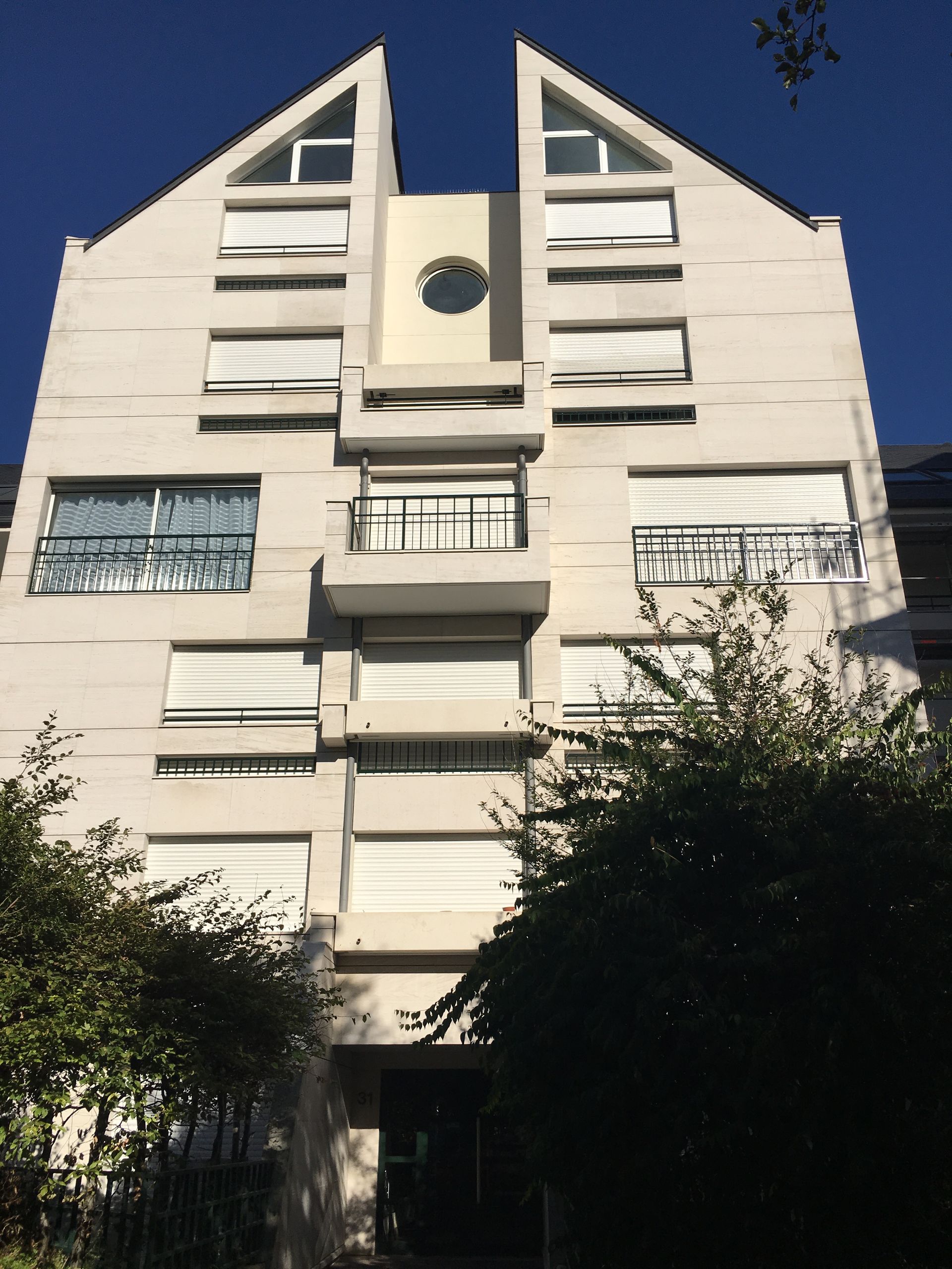 Immeuble blanc avec deux sommets en forme de triangle, des fenêtres et des stores fermés sous un ciel bleu.