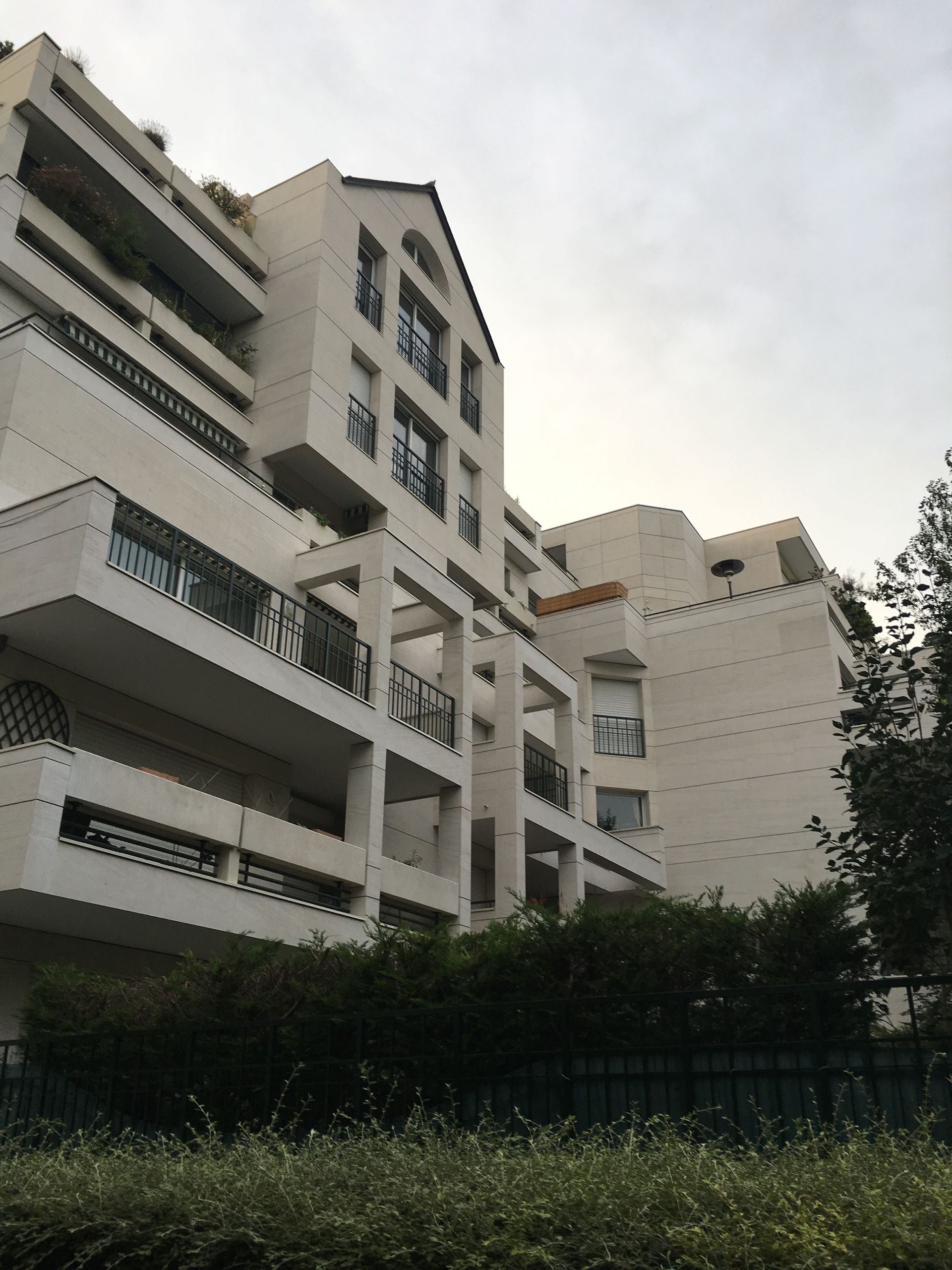 Immeuble d'appartements blanc à plusieurs étages avec balcons et verdure devant.