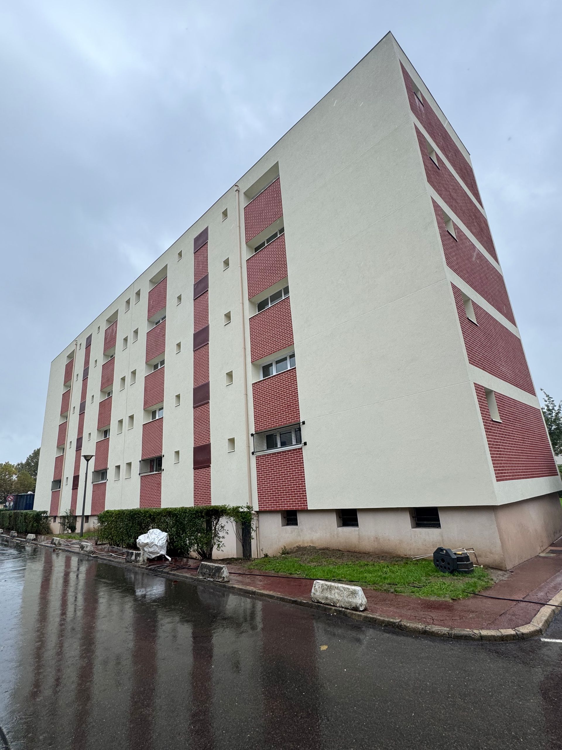 Immeuble d'appartements à plusieurs étages avec des murs beiges, des balcons à rayures rouges et une rue pluvieuse.