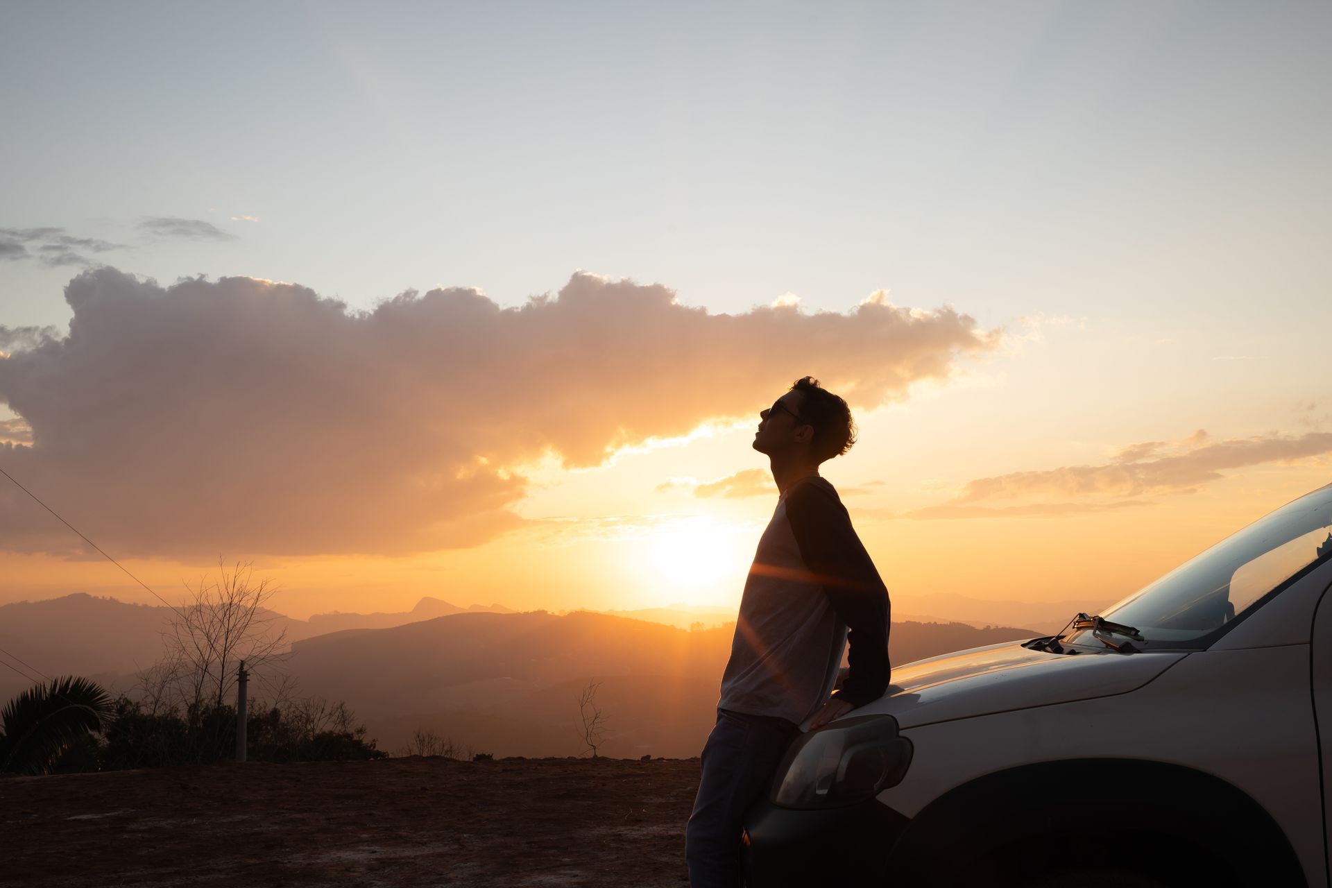 Un homme appuyé sur une voiture regarde le coucher de soleil sur les montagnes.