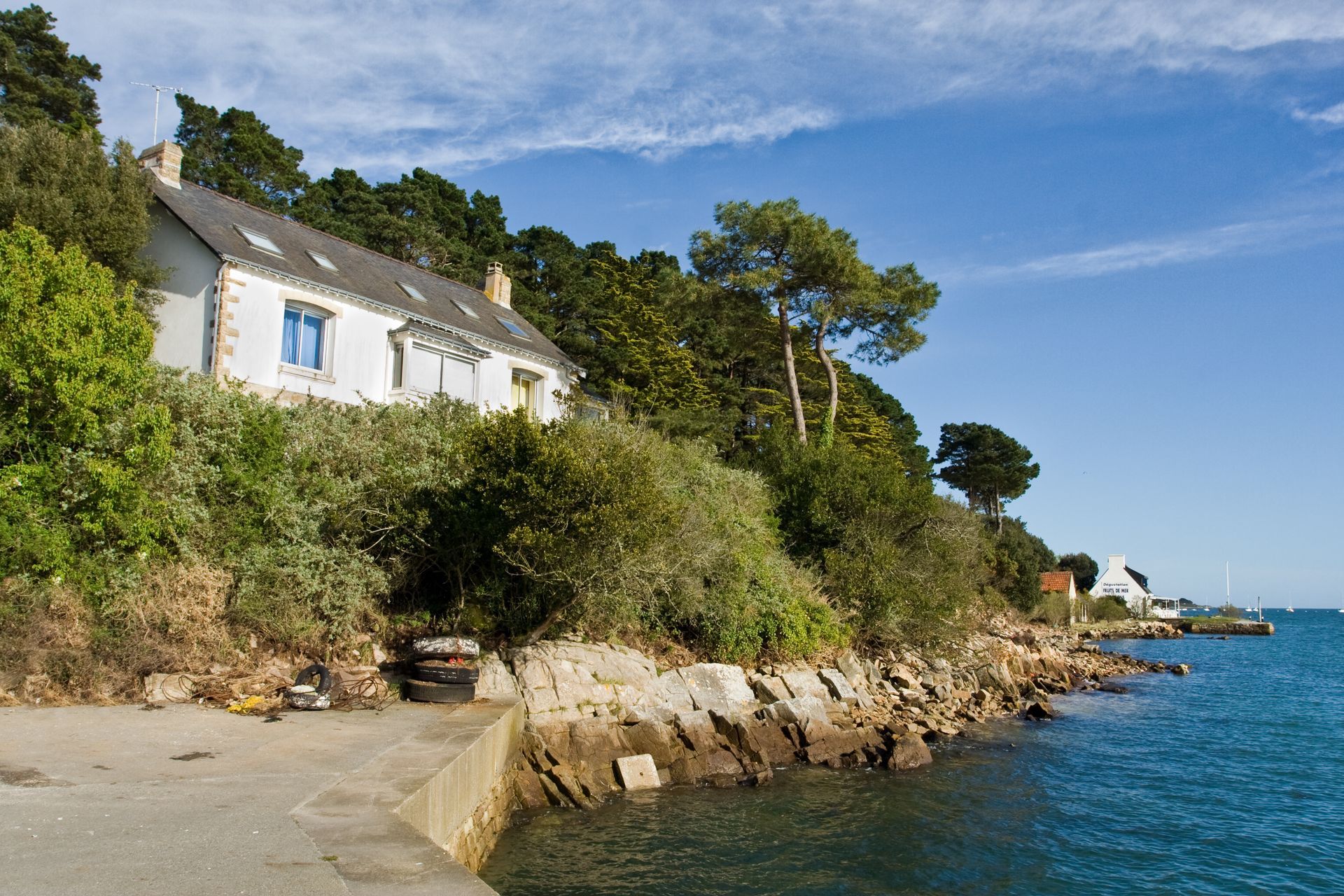 Maison blanche sur une côte rocheuse, avec eau et ciel bleus. Des arbres verts l'entourent.