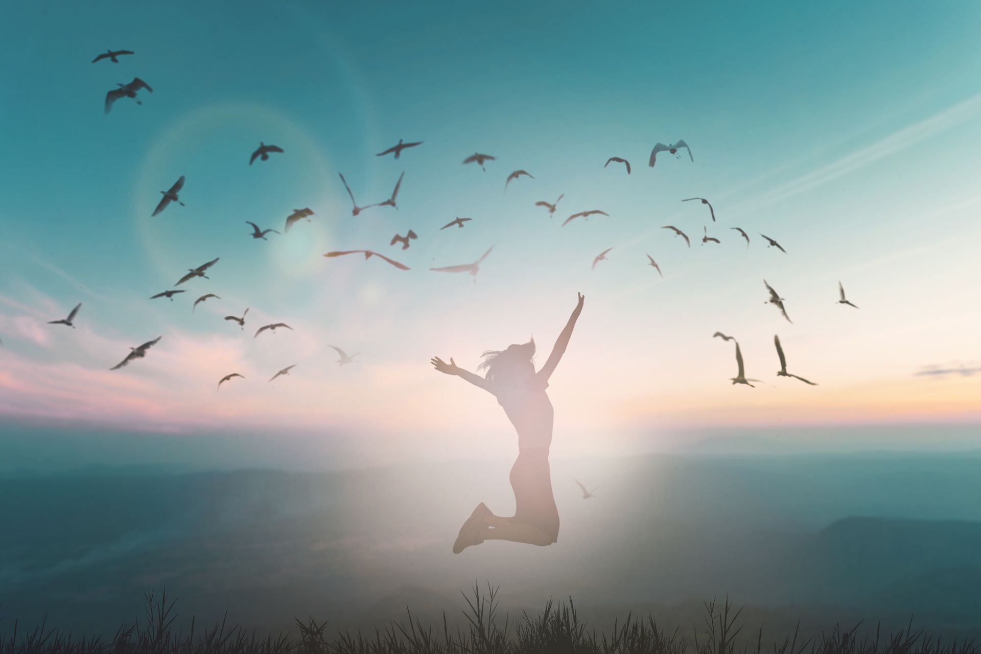 Silhouette d'une femme sautant les bras levés, oiseaux volant dans un ciel bleu sarcelle, coucher de soleil sur les montagnes.
