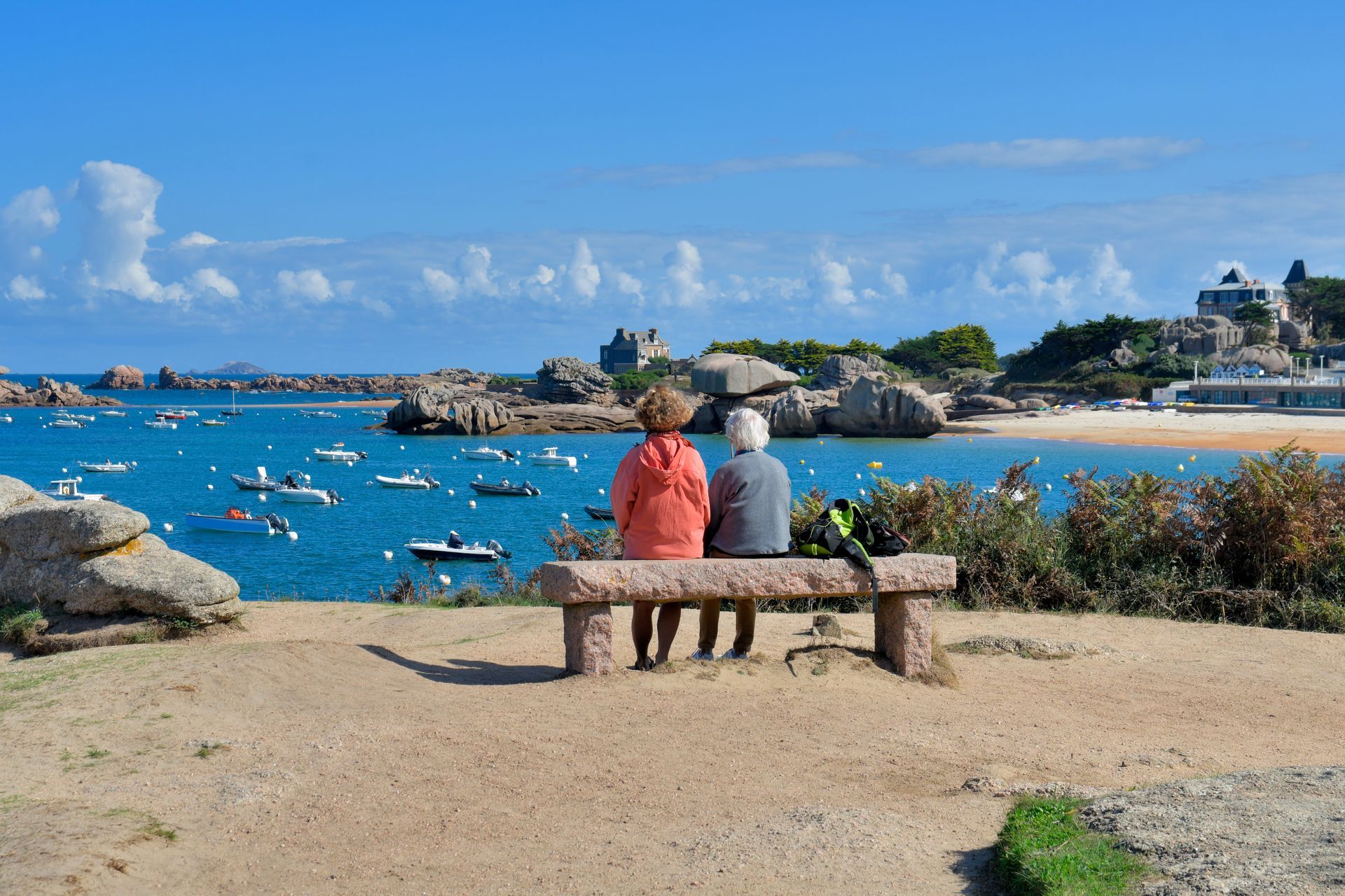 Deux personnes sont assises sur un banc surplombant une baie bleue parsemée de bateaux. Journée ensoleillée, rochers de granit rose.