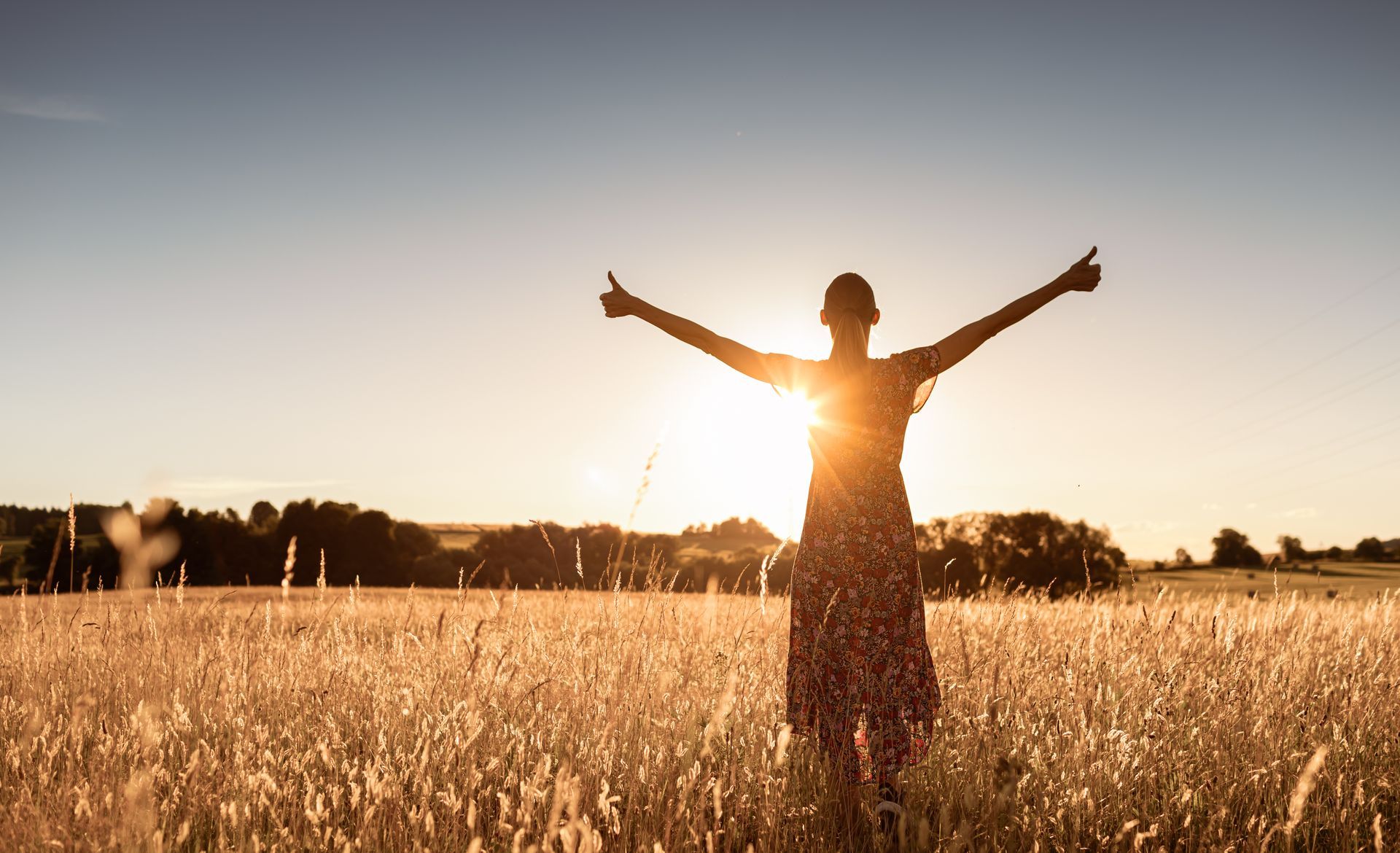 Femme debout dans un champ, les bras levés vers le soleil, se découpant dans un coucher de soleil doré.