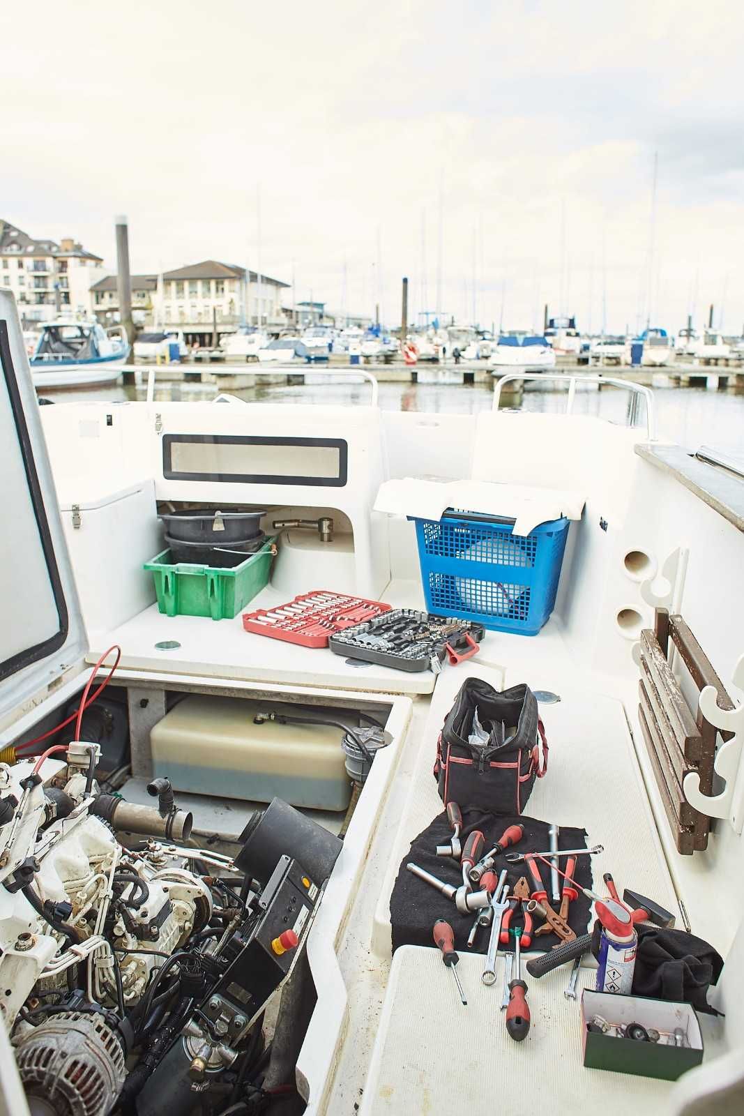 Cubierta de barco con compartimento del motor, bolsas de herramientas y barcos de puerto deportivo al fondo.