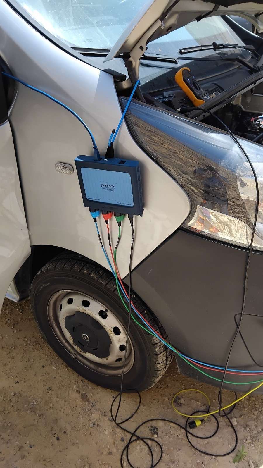 Dispositivo de diagnóstico azul sujeto al lateral de un coche blanco con cables y herramientas en el suelo.