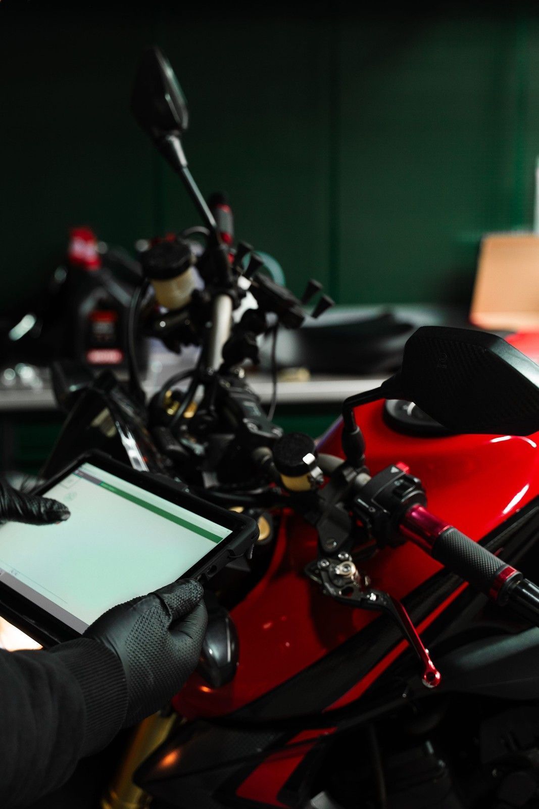 Manos enguantadas usando una tableta junto a una motocicleta roja en un garaje con poca luz.