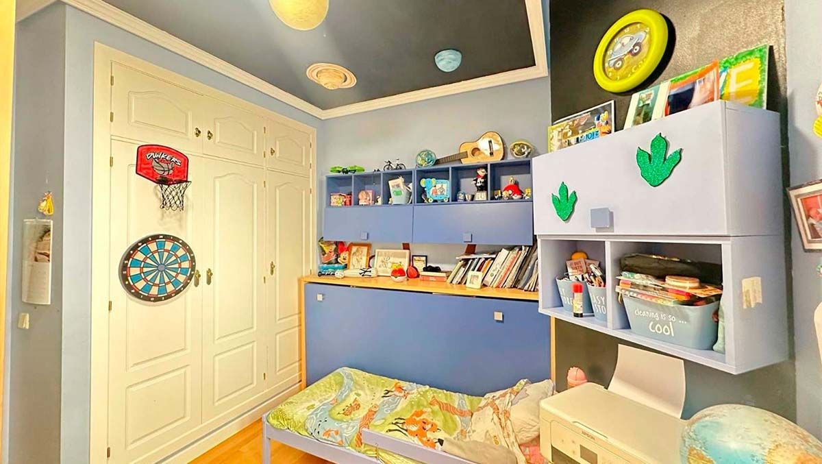Dormitorio infantil azul con cama, escritorio, estantes llenos de juguetes y una ventana.