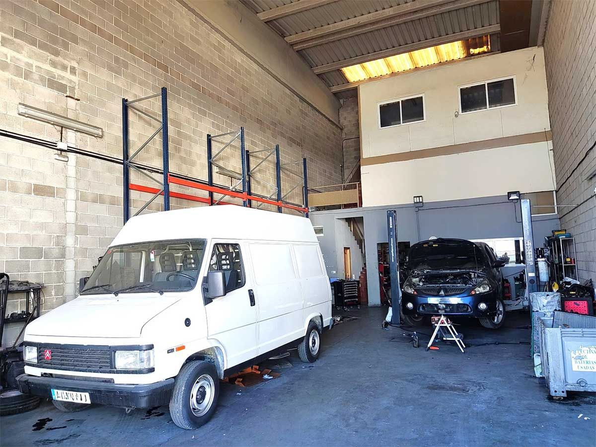 Furgoneta blanca y coche negro en el taller de un mecánico; una estructura de dos pisos en la parte trasera.