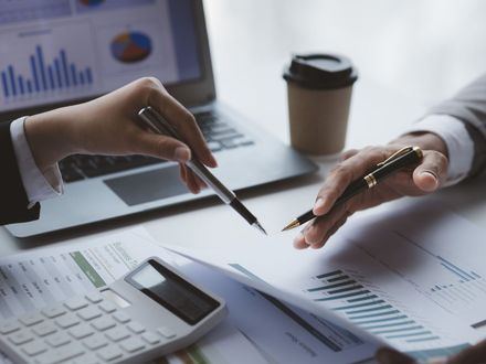 Dos personas revisando documentos financieros con bolígrafos, computadora portátil, calculadora y café.