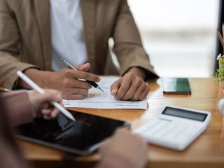 Dos personas revisan documentos en un escritorio. Una apunta con un bolígrafo y la otra usa una tableta. Hay calculadora y teléfono.
