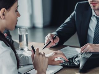 Mujer y hombre en traje de negocios en un escritorio, revisando papeles y usando una calculadora.