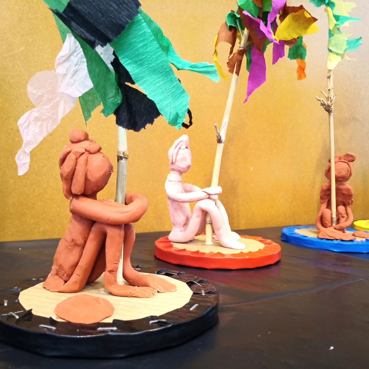 Esculturas de arcilla de personas sentadas bajo sombrillas sobre una mesa.