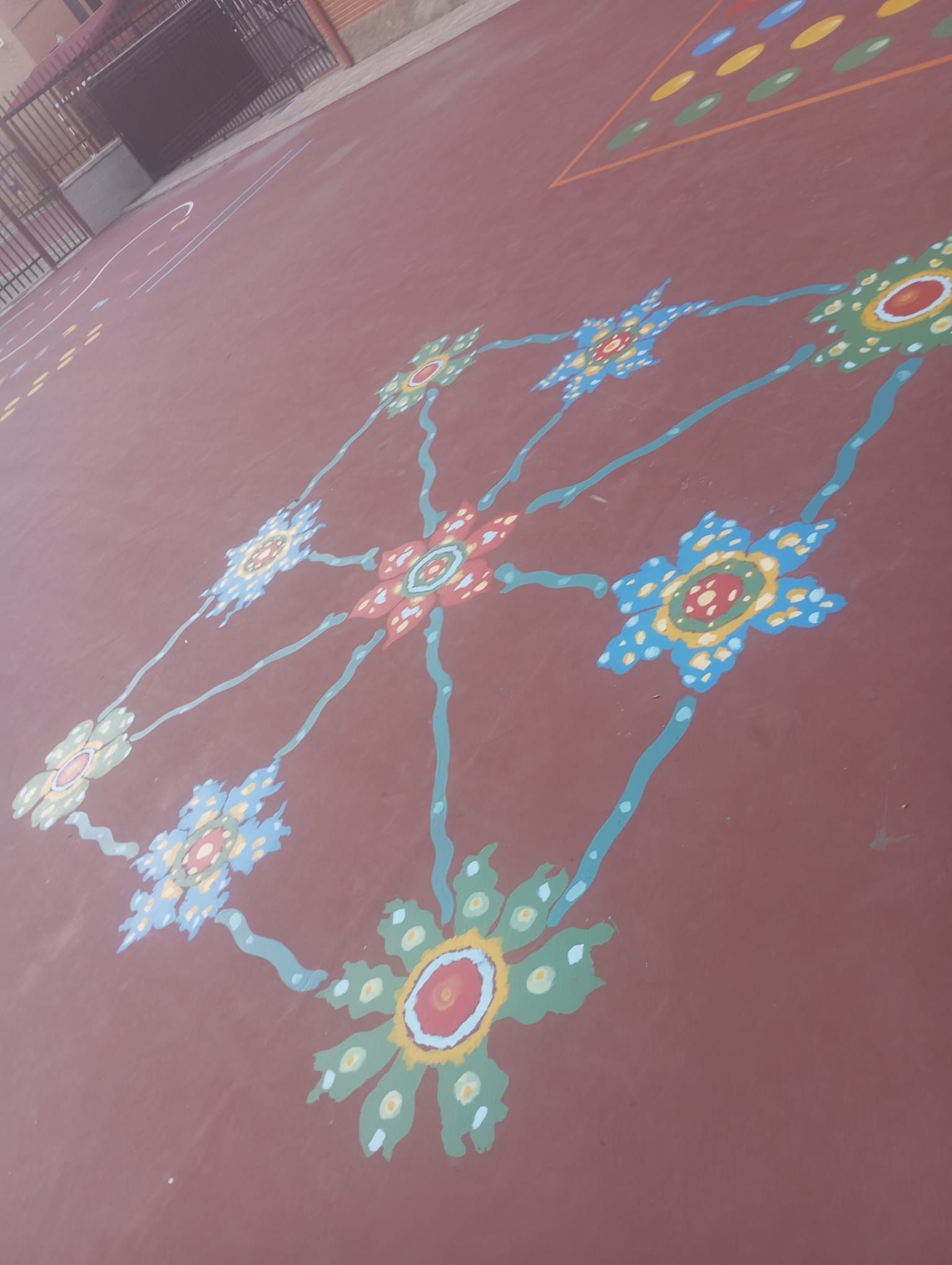 Diseño floral de rangoli colorido sobre un suelo de patio rojizo con una pared de ladrillos al fondo.