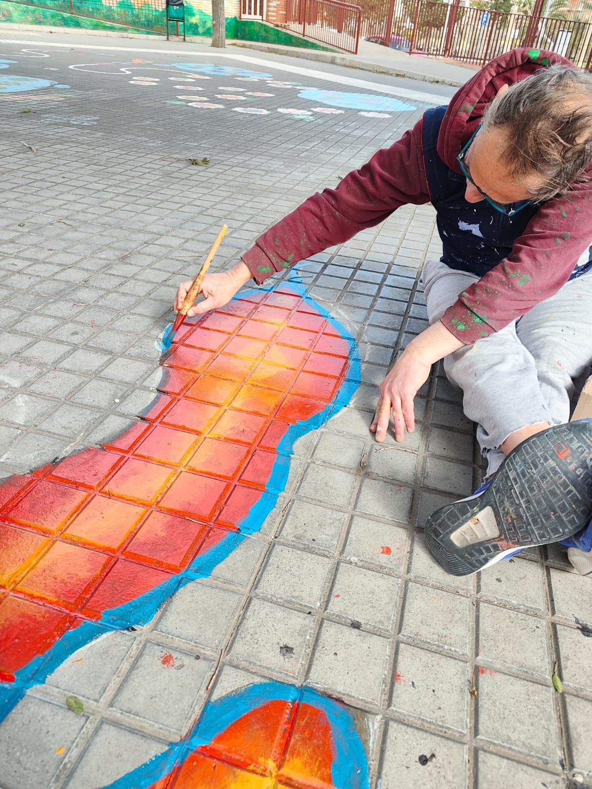 Persona pintando un mural rojo anaranjado sobre baldosas de la acera con un contorno azul.