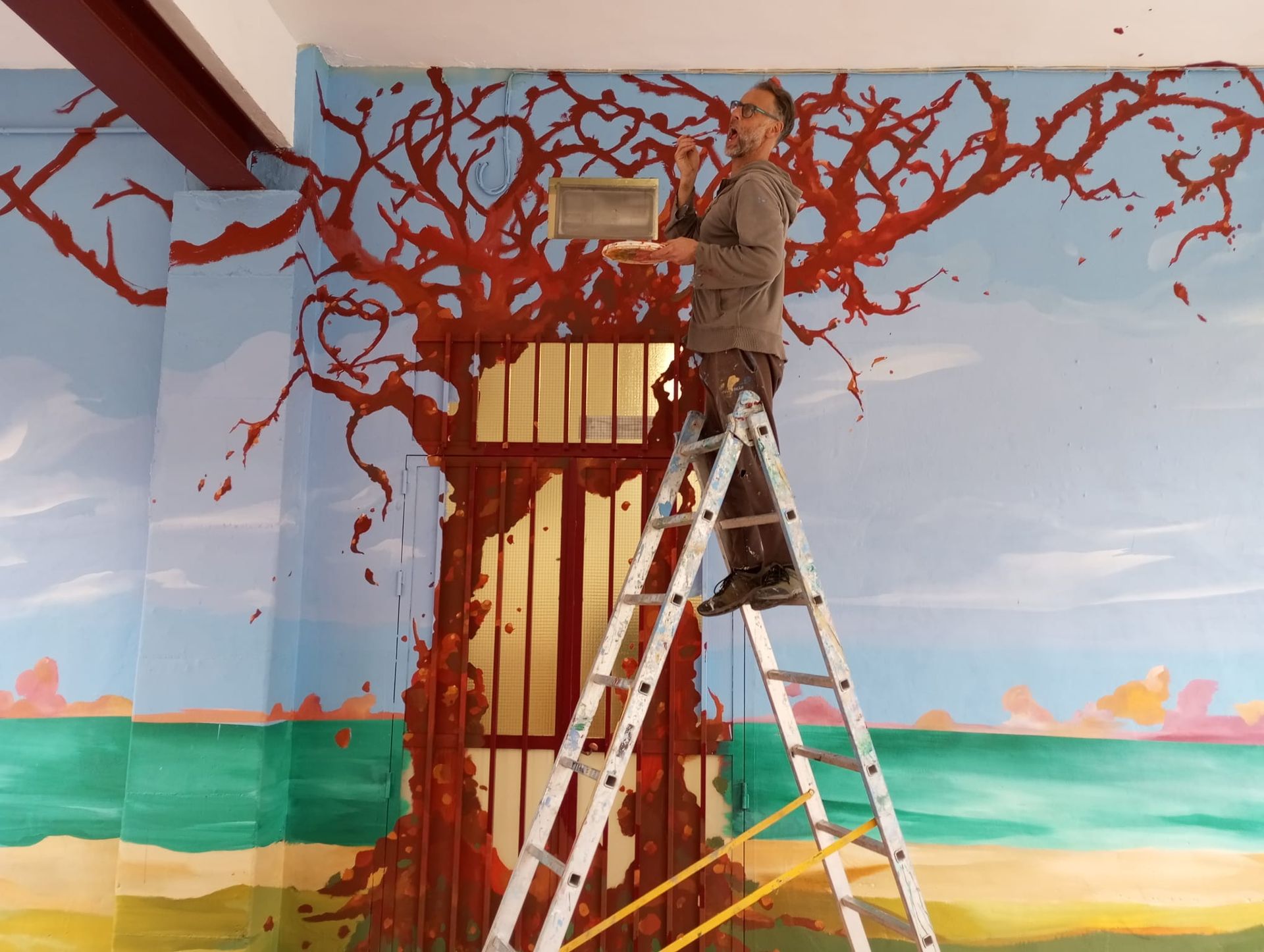 Una persona subida a una escalera pinta un mural de un árbol con ramas rojas en la pared de una habitación colorida.