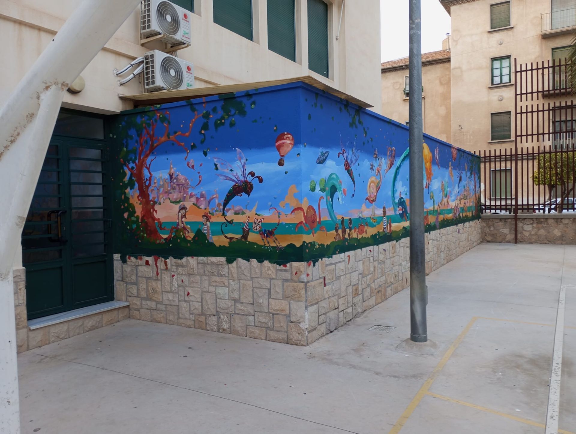 Patio exterior con un mural del océano azul en la pared de un edificio, base de piedra y pasarela pavimentada.