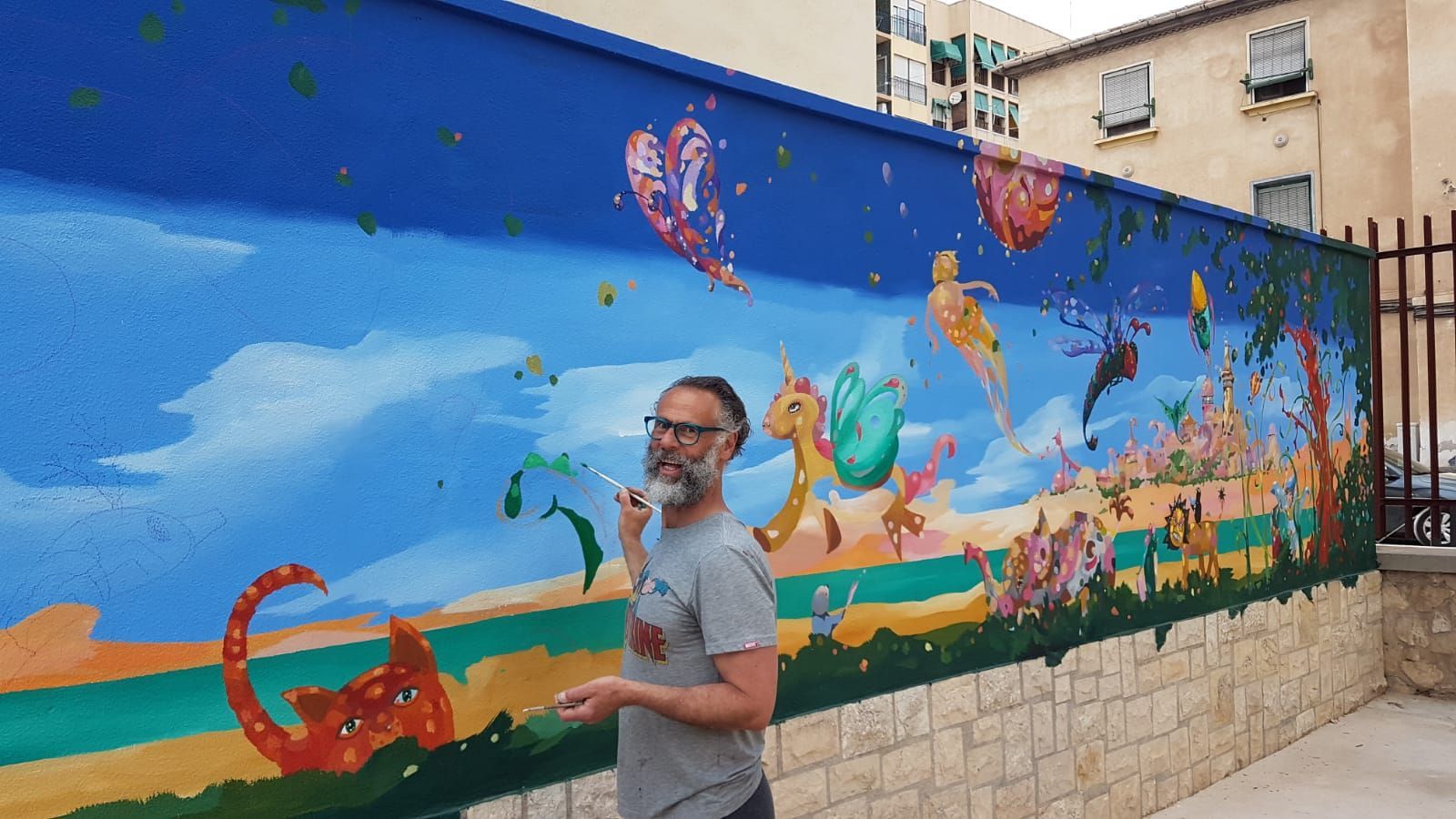 Un hombre de pie frente a un colorido mural de criaturas marinas y el cielo en una pared exterior.