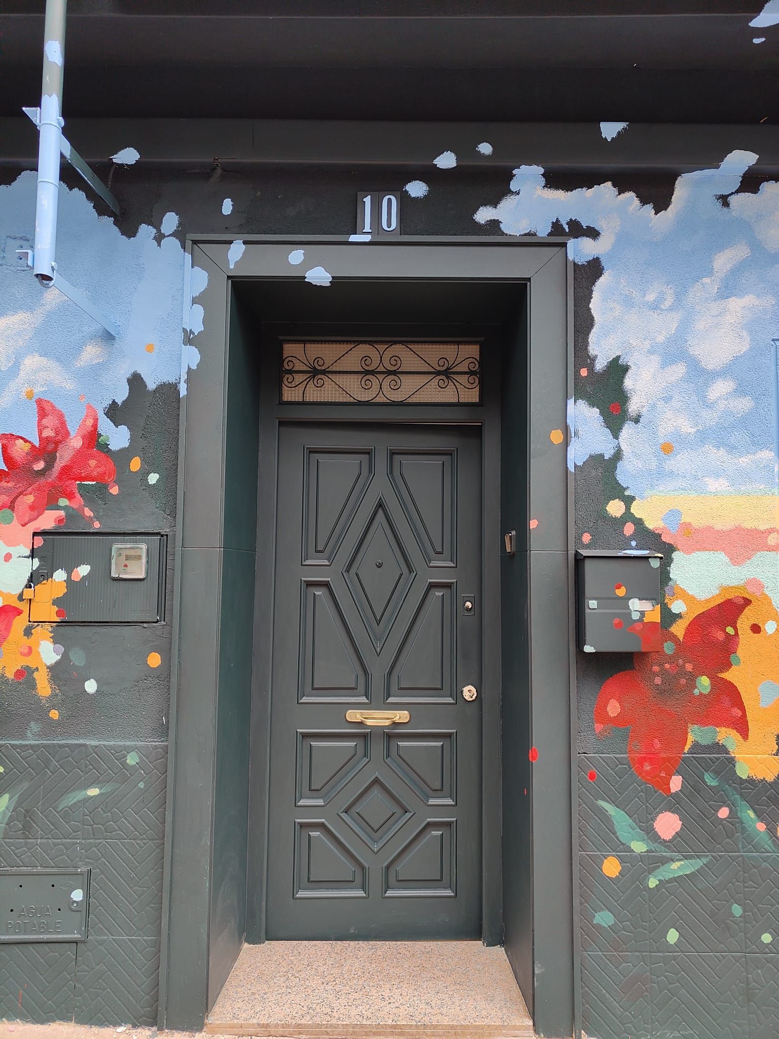 Puerta negra con paneles geométricos debajo del número 10, enmarcada por un colorido mural floral.