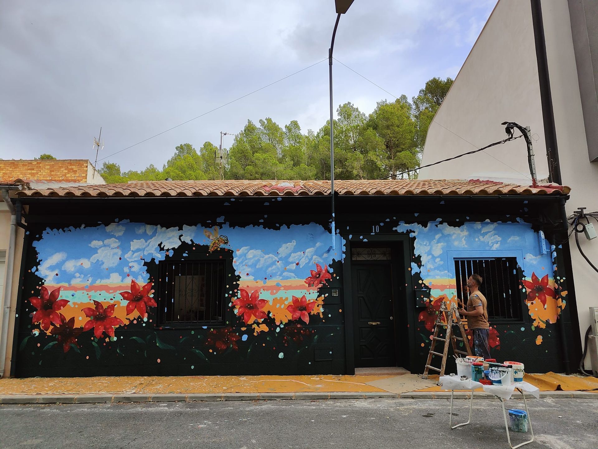 Mural colorido en la fachada de un pequeño edificio, con una puerta, una ventana y una persona de pie junto a la entrada.